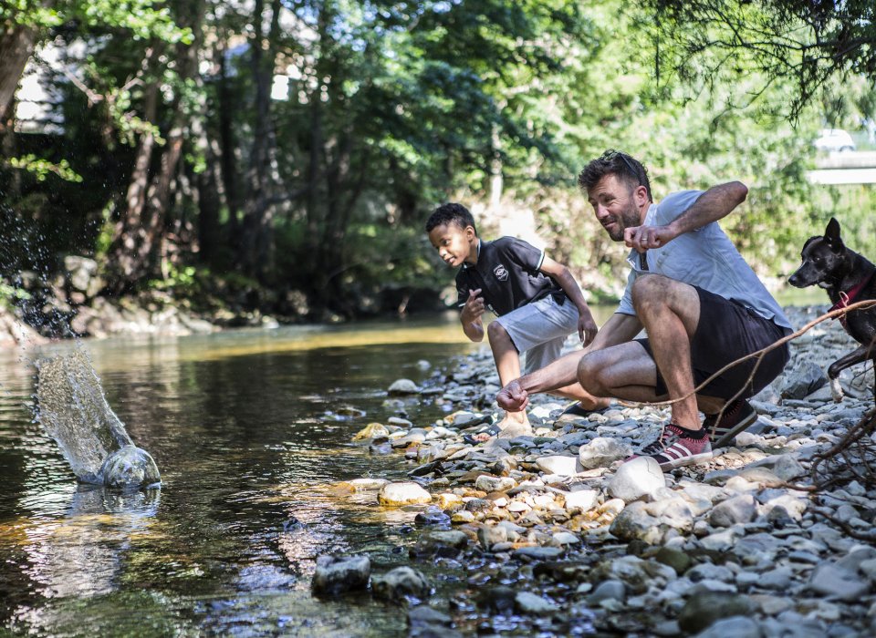 Un homme et un petit garçon jouent dans la rivière et font des ricochets. Un chien les observe derrière.